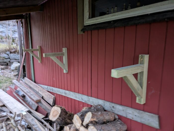 Träkonsoler fästa på en friggebods vägg för att lagra spillvirke; ved och plankor syns på marken intill.