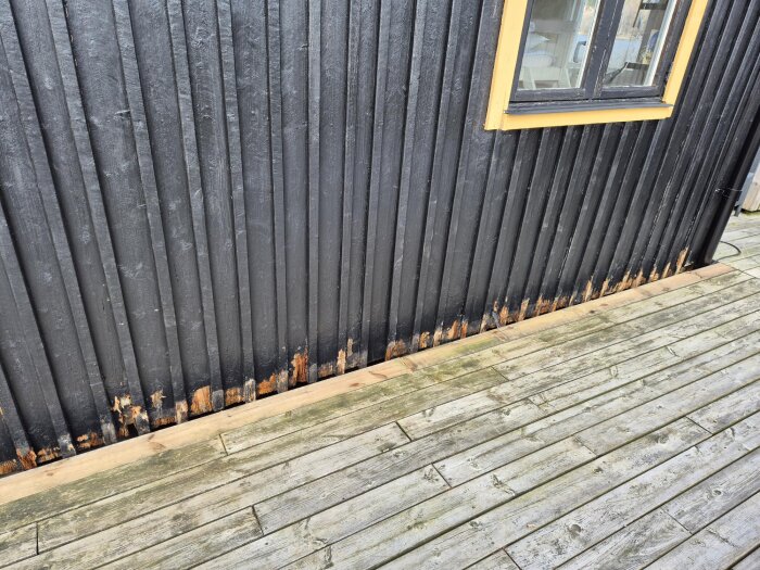 Närbild av en altan vid en svartmålad fasad med avskrapad färg nederst på brädorna, vid ett hus med träsidor och gult fönster.