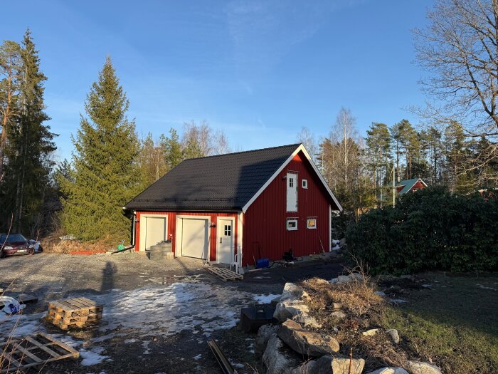 Rött garage med svart tak omgiven av vårträd och stenar, med träpallar och bil i förgrunden. Solig dag med klar himmel.