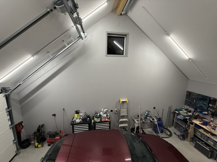 Garage med billyft, fritt golv och verktygsförvaring. Väggar med ljusrör och tak med garageöppnare. Stege och arbetsbänk synliga.