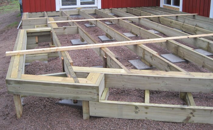 Bjälklag av trä för trädäck, uppbyggd på regelkonstruktion med grusunderlag, vid röd husfasad.