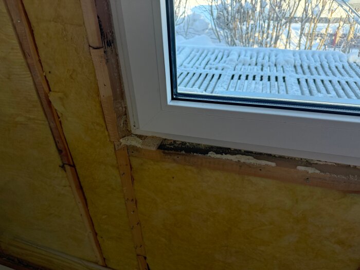 Fönster med ojämn spalt till regel, omgiven av isolering, visar potentiella problem med fogning och konstruktion. Snöig utsikt syns genom fönstret.