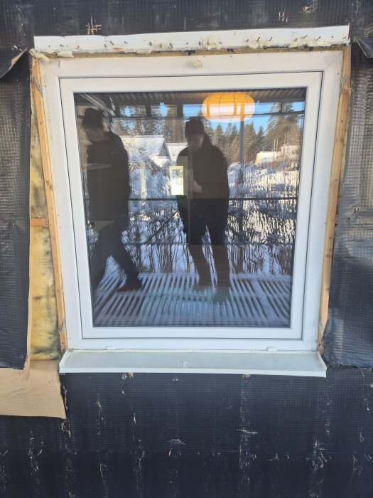 Ett nyrenoverat fönster på en svart fasad med liten spalt mellan fönsterkarm och regel, reflektion av två personer i glaset.