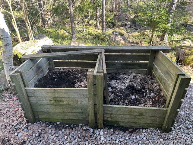 Två träkomposter utomhus i en skogsmiljö, fyllda med olika stadier av nedbrutet material, eventuellt för latrin- eller trädgårdskompostering.