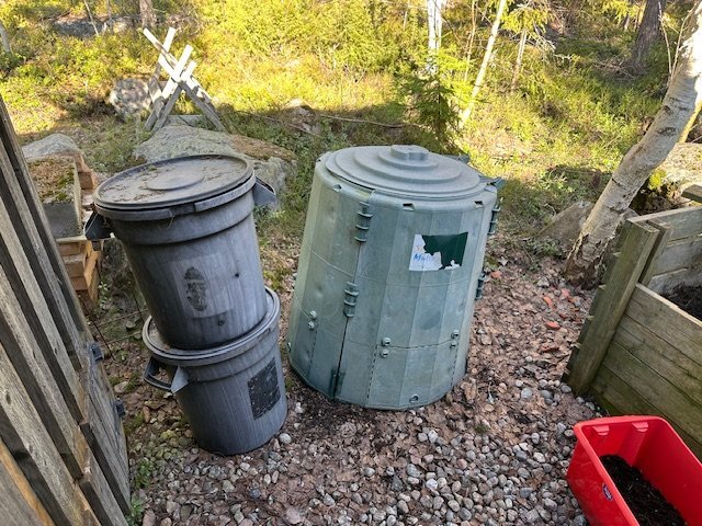 Två kompostkärl bredvid ett större grönt plastkärl på en grusad yta, omgiven av träd och skog. Möjligen del av en latrinkompost.