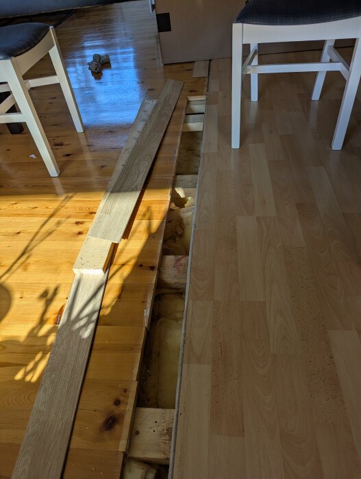Upprivet golv med synliga bjälkar och isolering, två vita stolar runtom, pågående golvrenovering.