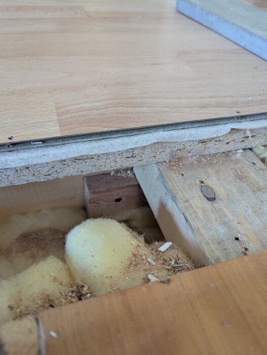 Uppbrutet laminatgolv visar träbjälklag och isolering av gul mineralull.