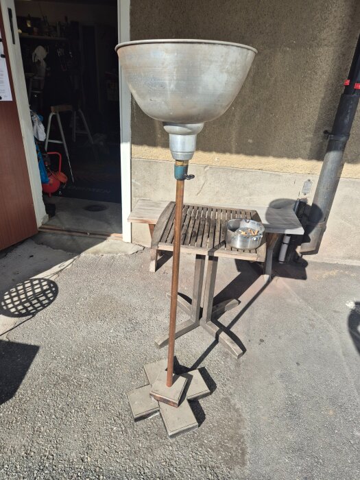 Hög metalltrattmontering på en stång med betongplattor som bas, placerad utomhus bredvid ett bord och en byggnad.