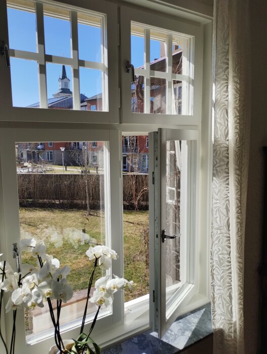 Öppet fönster med vita spröjs mot en trädgård och hus, orkidée på fönsterbrädan, soligt väder, kyrktorn syns i bakgrunden.