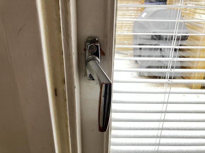 Handtag på en dörr med persienner i fönstret, där utomhus omgivning syns svagt genom glaset.