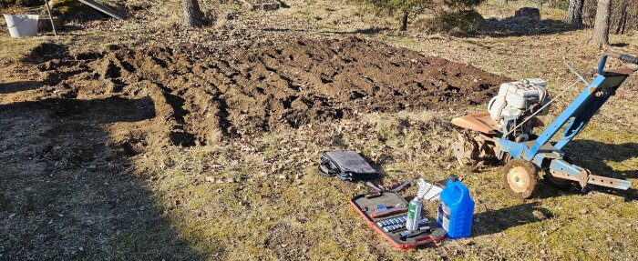Fräst jord i trädgård med en jordfräs och verktyg liggande på marken, inklusive en blå dunk och verktygslåda.
