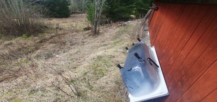 Röd vägg av carport på landsbygd, omgiven av torrt gräs och träd; blå plastkanister lutad mot väggen.