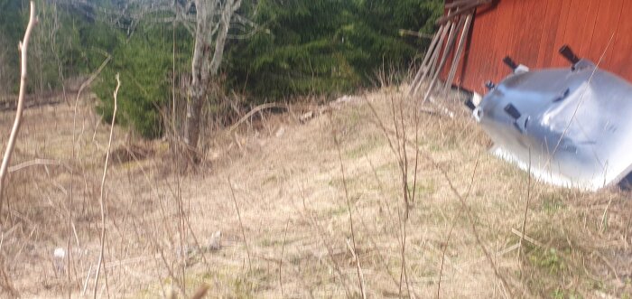 Brant sluttning bakom en röd byggnad med träd och gräs. En tank ligger bredvid byggnaden, delvis dolt av vegetationen.