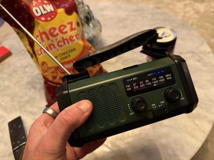 Hand håller en grön radio med antenn, en påse OLW Cheez Cruncherz och en burk dryck syns i bakgrunden på ett bord.