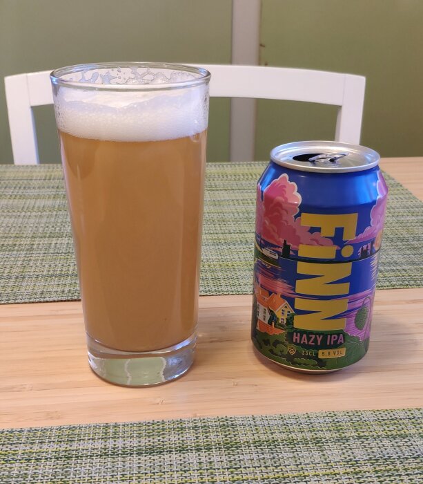 Glas med ljus, disig öl bredvid en färgglad burk märkt "Finn Hazy IPA", placerad på ett bord med grönmönstrad duk.