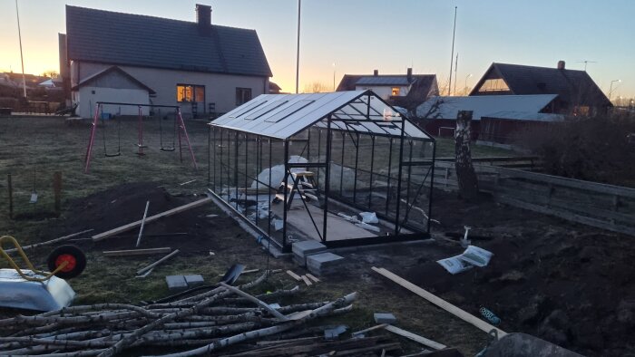Växthus under konstruktion vid solnedgång, omgiven av verktyg och brädor i en trädgård med lekplats i bakgrunden.