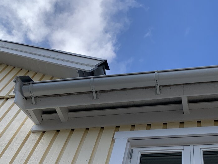 Vindskivor och takränna på gult hus, blå himmel i bakgrunden. Nyinstallerade plankor, del av diskussion om byggkvalitet.