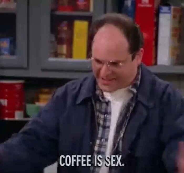 En man i flanellskjorta och glasögon står i köket med en text på skärmen: "COFFEE IS SEX".