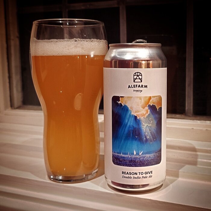 Ett glas med gyllene öl bredvid en ölburk från Alefarm Brewing med etiketten "Reason to Give" på ett vitt fönsterbräde.