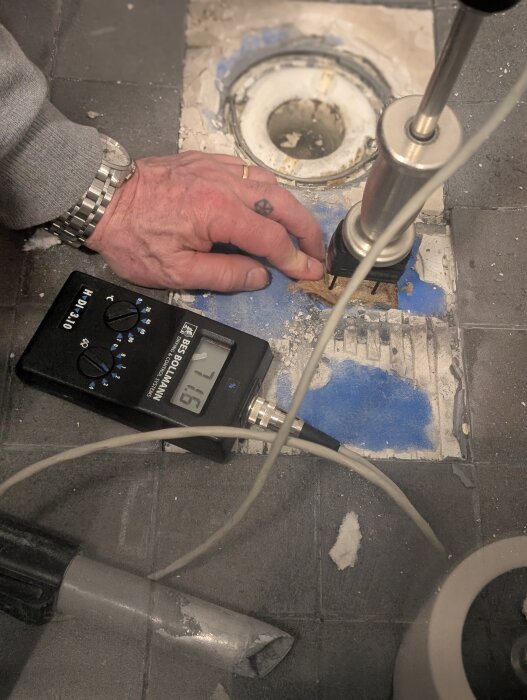 Hand mäter fukt i ett badrumsgolv med en fuktmätare nära ett avlopp. Verktyg och tråd syns på mörka kakelplattor.