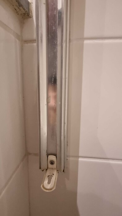 Metallfäste på kaklad vägg med skena, skruvhål synligt, del av okänt objekt i bakgrunden. Används tidigare för glasvägg.
