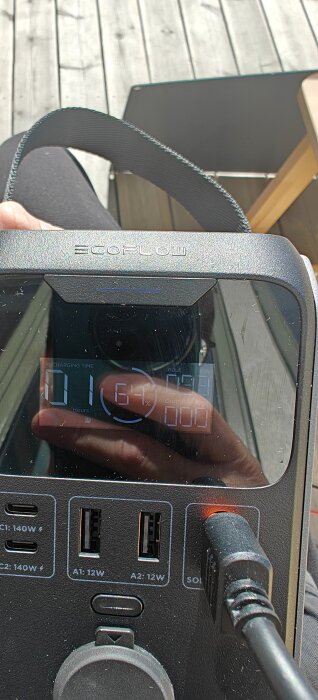 Bärbar EcoFlow-powerbank med display som visar laddningstid, anslutna kablar och USB-portar, fotad på en veranda med trallgolv.