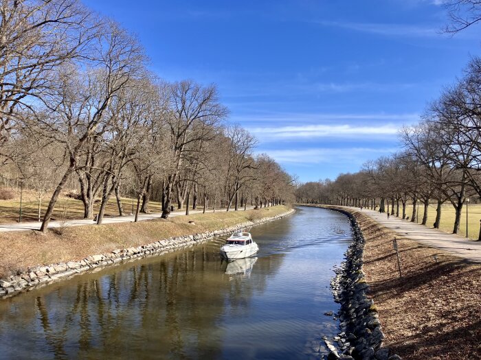 Båt på en lugn kanal omgiven av träd utan löv, en gångväg på båda sidor. Klar blå himmel över en vår- eller höstdag.