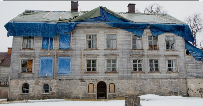Stort äldre hus med trasigt tak täckt av blå och gröna presenningar, många fönster, sliten fasad och grå stenbas.
