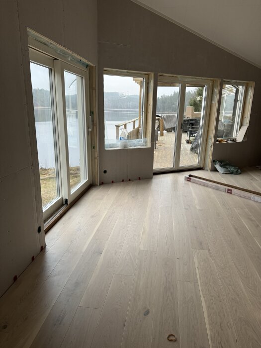 Nylagt trägolv i ett rum med stora fönster och utsikt över en sjö och skog, byggmaterial syns utanför på en veranda.