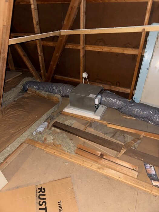 Ett vindsutrymme med synliga träbalkar, isoleringsmaterial och ventilationsrör, samt kartong och verktyg på golvet.