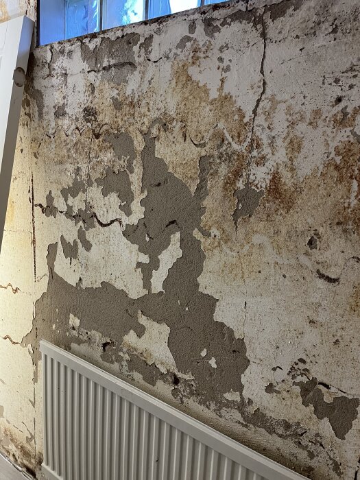 Källarvägg med avflagnad färg och sprickor, panel borttagen, synlig radiator.