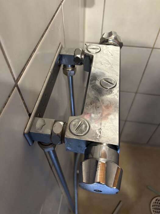 Kromad badrumstermostat monterad på vitkaklad vägg, med synliga anslutningar och vred.