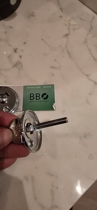Förzinkad ASSA vredmedbringare hålls i handen, med en Beslagsboden-förpackning synlig i bakgrunden.