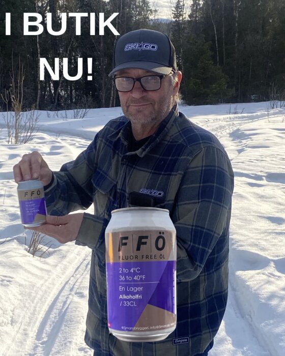 Man i keps och flanellskjorta håller en burk alkoholfri öl med texten "I BUTIK NU!", snötäckt bakgrund.