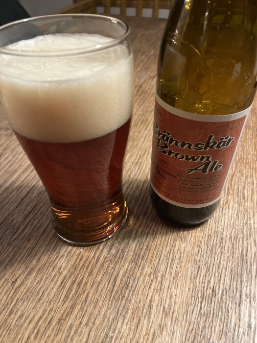 Ett glas fyllt med brun ale med skum, intill en flaska märkt “Brännskär Brown Ale” på ett träbord.