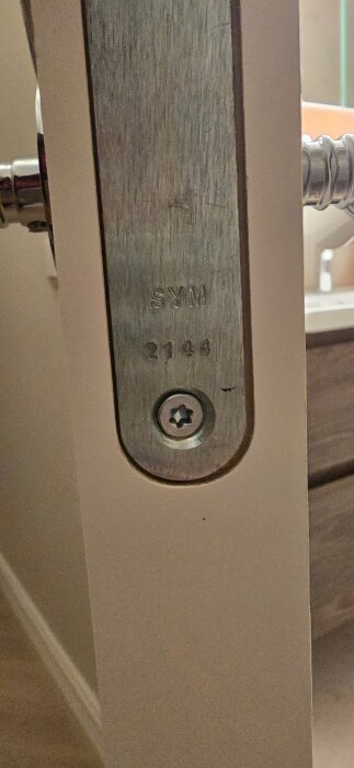 Närbild på metallplatta på en dörr med texten "SYN 2144" och en synlig stjärnformad skruv.