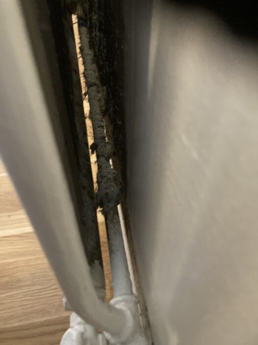 Gammelt varmeelementrør bak radiator med lag av beige materiale, potensielt gammelt isolasjonsmateriale.