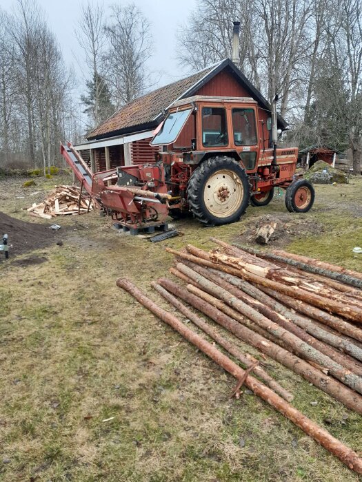Traktor med vedklyvningsmaskin framför en lada, omgiven av staplade trästammar och vinterträd i bakgrunden.
