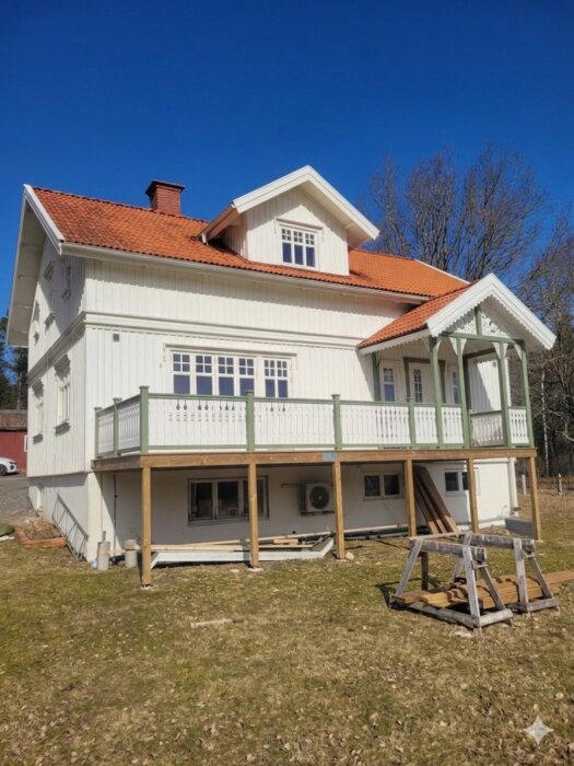 Vit tvåvåningshus med röd tegelvilla och pågående altanbygge. Träräcke planeras till sommaren, farstukvist nästa år.