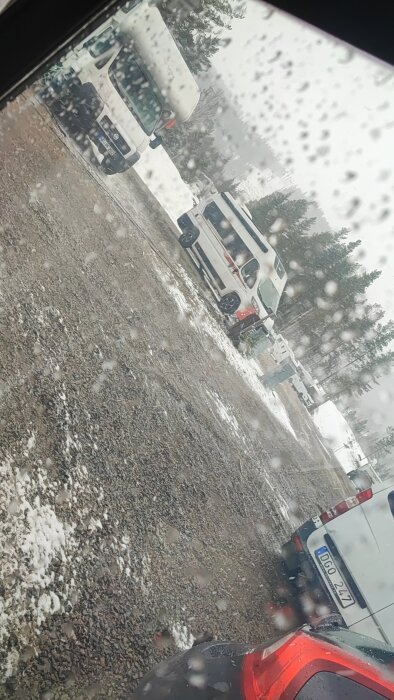Bilar parkerade på en snötäckt och regnig plats i skogen, fotat genom ett fönster med vattendroppar.