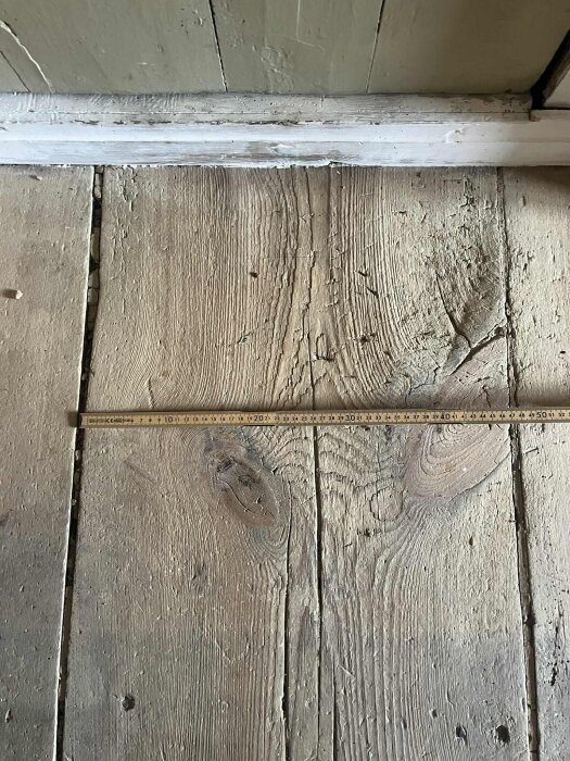 Gammalt trägolv i ett 1800-talshus med slitage, uppstickande spetor och en mätskala för storleksreferens.
