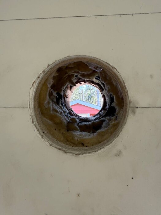 Runt hål i innervägg med isolering och utsikt över en röd byggnad utanför, förberett för installation av friskluftsventil.