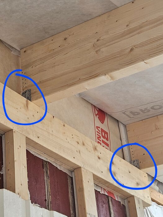 Träkonstruktion med synliga reglar och metalldelar, två blå markeringar vid metallbeslag som fäster reglar i tak. Byggmaterial i bakgrunden.