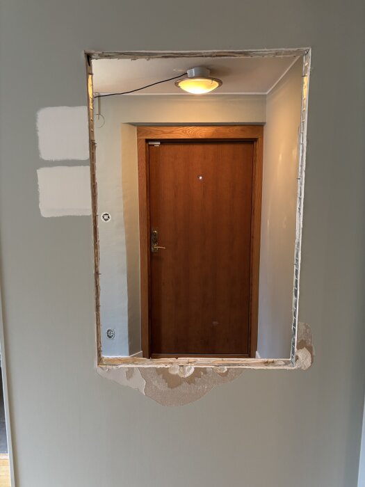 Öppning i vägg efter borttaget fönster, omlackerad och skarvspacklat, bredvid en trädörr och under en plafondlampa.