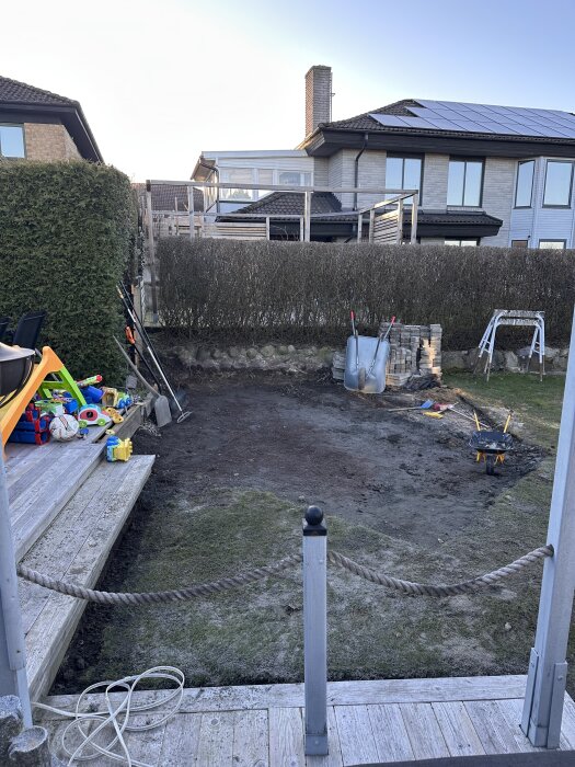 Trädgård med hårt packad jordyta, leksaker, skottkärra och vedtrave, planeras för gräsmatta istället för altan.