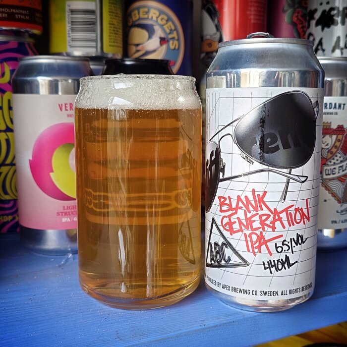 Ett glas ljus öl står bredvid en burk med etiketten "Blank Generation IPA" på en blå yta, omgivna av andra ölburkar.