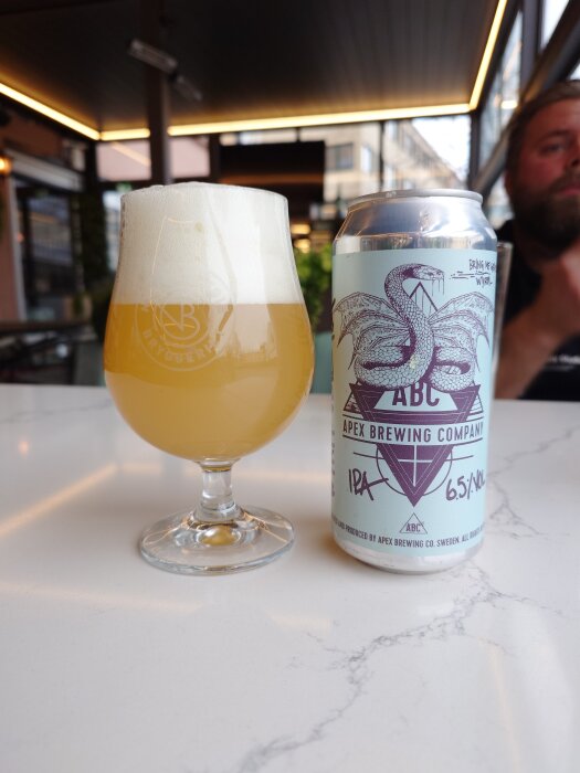 Ett glas med skummande öl och en burk från Apex Brewing Company på ett bord.