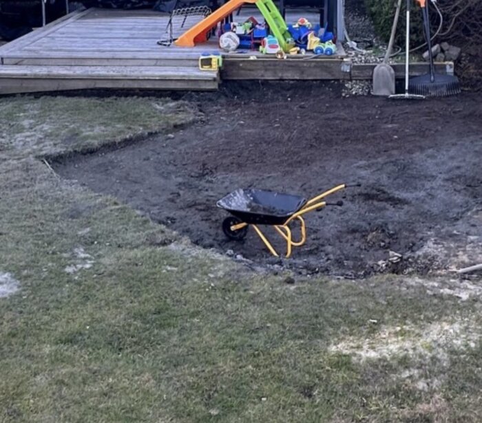Skottkärra på en jordig yta framför en veranda med leksaker och trädgårdsredskap som kratta och spade intill.