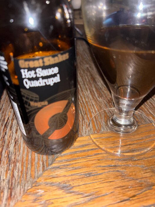 Flaska märkt "Hot Sauce Quadrupel" och ett glas med mörk dryck på ett träbord.