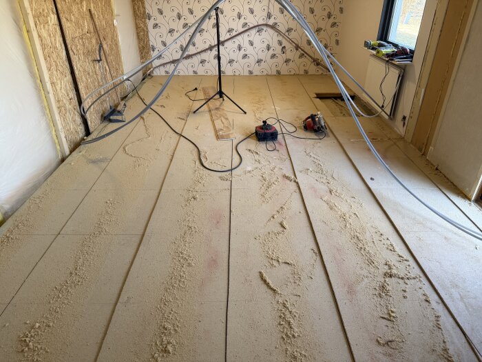 Rivet golv med träspån och kablar, verktyg på golvet, tapetserad vägg i bakgrunden, renoveringsprojekt i ett rum under arbete.